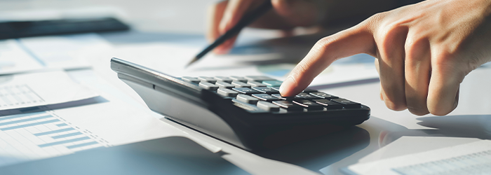 Hand clicking button on calculator while other hand is holding pen to financial papers in background