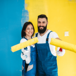 Man and woman standing together holding paint rollers with half yellow half teal wall behind them