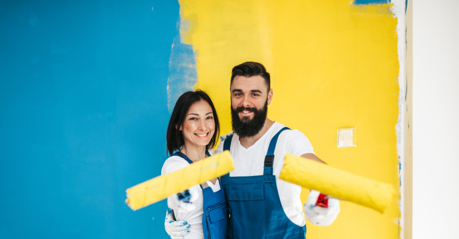 Man and woman standing together holding paint rollers with half yellow half teal wall behind them